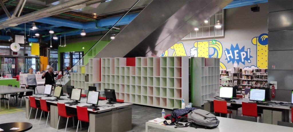 Estantería escalonada roja, blanca y verde bajo la escalera mecánica de la biblioteca