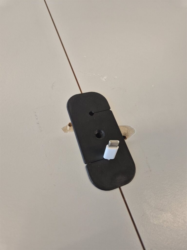 Black 3D-printed USB cable holder fixed over the joint between two table tops.